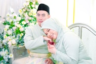 A couple dressed in matching light green attire, with the woman resting her head on the man's shoulder. The woman's pose suggests affection, and her hand features intricate henna designs. The backdrop is adorned with an abundance of white and yellow flowers, adding to the festive and celebratory atmosphere.