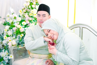 A couple dressed in matching light green attire, with the woman resting her head on the man's shoulder. The woman's pose suggests affection, and her hand features intricate henna designs. The backdrop is adorned with an abundance of white and yellow flowers, adding to the festive and celebratory atmosphere.