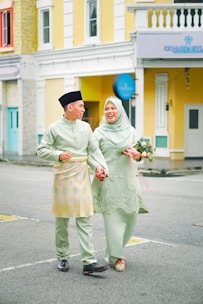 A joyful couple walking hand in hand, dressed in matching traditional attire. The woman holds a bouquet of flowers, and both individuals are smiling warmly, conveying a sense of happiness. The background features colorful buildings with a mix of yellow, blue, and white facades.
