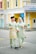 A joyful couple walking hand in hand, dressed in matching traditional attire. The woman holds a bouquet of flowers, and both individuals are smiling warmly, conveying a sense of happiness. The background features colorful buildings with a mix of yellow, blue, and white facades.