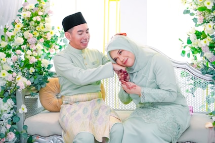 A young Muslim couple smiling warmly, the woman wearing a hijab and the man with a beard, sitting together with love symbols softly glowing around them.