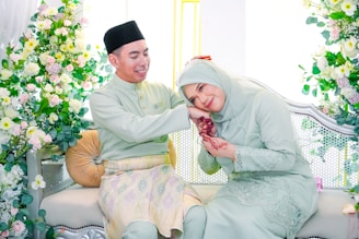 A couple dressed in traditional attire sits on an elegant sofa adorned with floral decorations. The woman, wearing a hijab and a pastel-colored dress, leans her head affectionately on the man's shoulder with henna on her hands. The man smiles warmly, wearing a matching outfit and a black hat. Vibrant flowers in pink, white, and green surround them, creating a celebratory and intimate atmosphere.