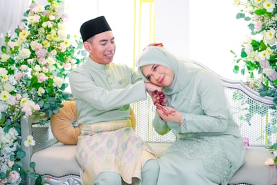 A couple dressed in traditional attire sits on an elegant sofa adorned with floral decorations. The woman, wearing a hijab and a pastel-colored dress, leans her head affectionately on the man's shoulder with henna on her hands. The man smiles warmly, wearing a matching outfit and a black hat. Vibrant flowers in pink, white, and green surround them, creating a celebratory and intimate atmosphere.