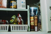 A pantry shelf filled with various packaged food items, including a large container of oats, a box of lactose-free product, and assorted snacks. Some items are organized in plastic baskets while others are stacked directly on the shelf.