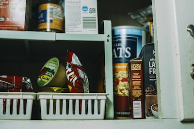 A pantry shelf filled with various packaged food items, including a large container of oats, a box of lactose-free product, and assorted snacks. Some items are organized in plastic baskets while others are stacked directly on the shelf.
