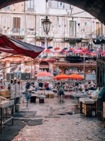 A bustling outdoor market scene with various stalls displaying goods under vibrant red umbrellas. Vendors and shoppers engage in lively interactions. The market is set in front of an old building with a rustic façade and balconies adorned with colorful pennants and flags. The cobblestone pathway glistens in the light, adding a lively ambiance to the scene.