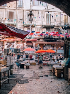 A bustling outdoor market scene with various stalls displaying goods under vibrant red umbrellas. Vendors and shoppers engage in lively interactions. The market is set in front of an old building with a rustic façade and balconies adorned with colorful pennants and flags. The cobblestone pathway glistens in the light, adding a lively ambiance to the scene.