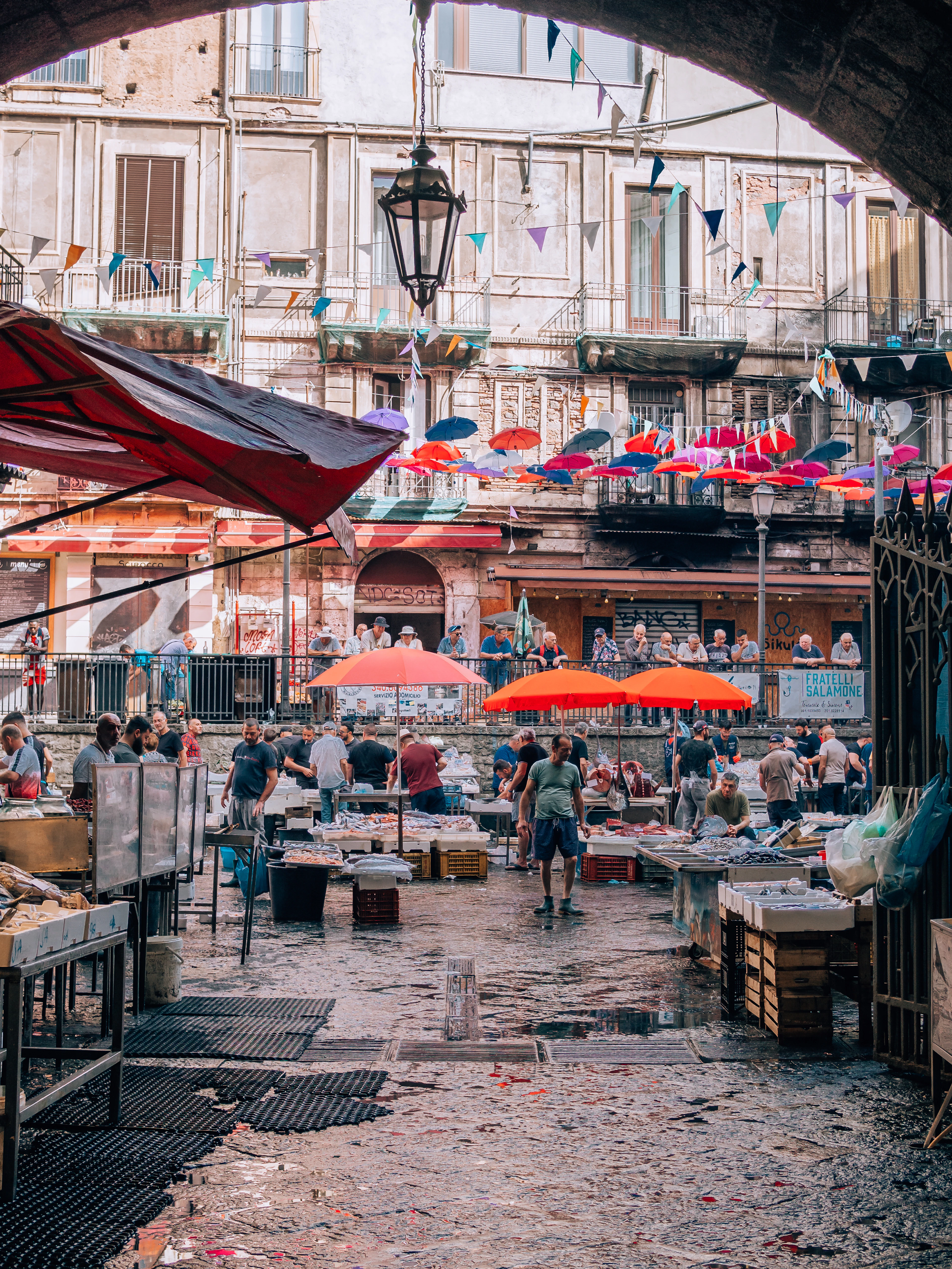 Catania's Pescheria fish market seen through the dark stone arch that leads into it, with wet lava paving, orange parasols and strings of bunting, trestles of iced fish and aproned fishmongers below a peeling baroque palazzo.