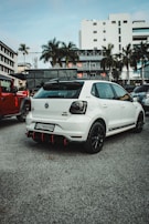 A white Volkswagen car with GT branding is parked on a concrete surface in an urban setting. The surroundings include buildings and palm trees, with several other parked vehicles visible. The car features a black roof rack and sporty black and red accents on the rear bumper.