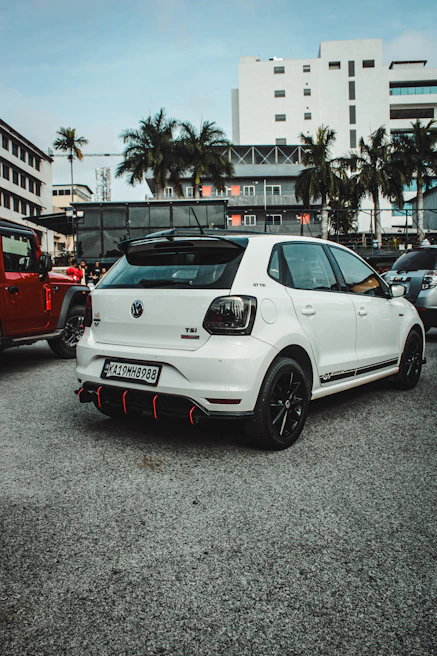 A white Volkswagen car with GT branding is parked on a concrete surface in an urban setting. The surroundings include buildings and palm trees, with several other parked vehicles visible. The car features a black roof rack and sporty black and red accents on the rear bumper.