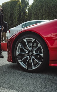 A close-up of a red car's wheel with sleek, modern alloy rims. The vehicle is parked on a textured surface with other cars visible in the background. There are several people standing nearby, and a hedge can be seen in the background, adding greenery to the scene.