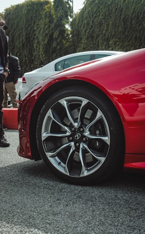 A close-up of a red car's wheel with sleek, modern alloy rims. The vehicle is parked on a textured surface with other cars visible in the background. There are several people standing nearby, and a hedge can be seen in the background, adding greenery to the scene.
