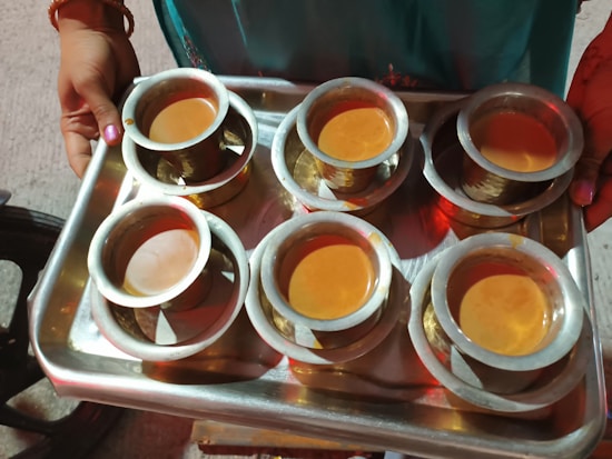 A metal tray holds six brass cups filled with a light brown liquid. The tray is being carried by a person whose hands have colorful bangles. The surroundings appear to be outdoors with a concrete surface visible.