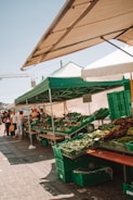 A bustling outdoor market stand with green and white tents offering various fresh vegetables in green crates. Several people are gathered around, browsing the produce, indicating a lively and busy atmosphere.