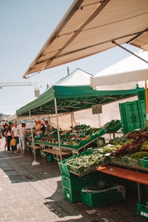 A bustling outdoor market stand with green and white tents offering various fresh vegetables in green crates. Several people are gathered around, browsing the produce, indicating a lively and busy atmosphere.
