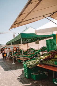 A bustling outdoor market stand with green and white tents offering various fresh vegetables in green crates. Several people are gathered around, browsing the produce, indicating a lively and busy atmosphere.
