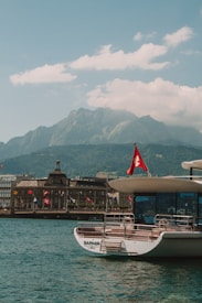 A serene waterfront scene with a boat named 'Saphir' moored in the foreground, flying a Swiss flag. The background features majestic mountains under a clear blue sky, accompanied by a few scattered clouds. Several buildings line the distant shore, also adorned with Swiss flags, creating a picturesque and calm setting.