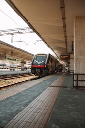 A train is parked at a station platform with tracks on either side. Several people are seen near the train, possibly boarding or alighting. Overhead power lines run above the train, and there is a clock and an electronic sign displaying train information. The station appears to be outdoors with natural light filtering through.