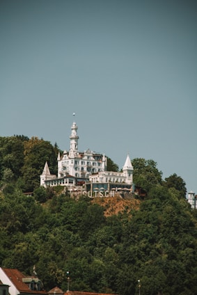A grand castle-like building with multiple turrets and spires sits atop a lush, forested hill. The structure is predominantly white with a mixture of architectural features ranging from pointed roofs to intricate balconies. Below the building, the large letters spell 'UTSCH', prominently displayed on the hillside.