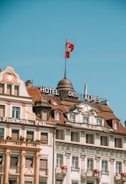 Historic European building facades with intricate architectural details and a Swiss flag waving atop one of the structures, set against a clear blue sky.