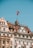 Historic European building facades with intricate architectural details and a Swiss flag waving atop one of the structures, set against a clear blue sky.