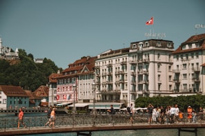 A scenic view of European-style buildings with ornate architecture, situated beside a calm body of water. A bridge crosses the water, where several people are walking. The scene includes lush greenery on a hill in the background, with a prominent sign on the hill. The Swiss flag flies atop one of the buildings.