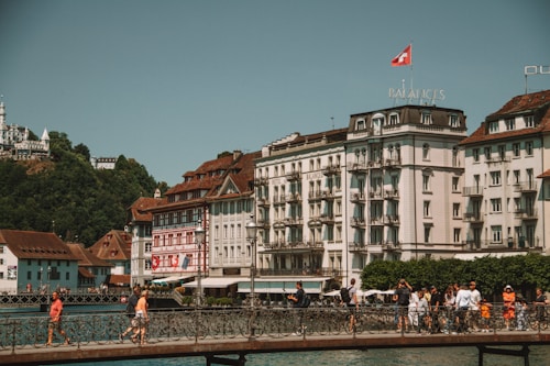 A scenic view of European-style buildings with ornate architecture, situated beside a calm body of water. A bridge crosses the water, where several people are walking. The scene includes lush greenery on a hill in the background, with a prominent sign on the hill. The Swiss flag flies atop one of the buildings.
