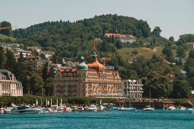 A grand building with distinctive architecture featuring an orange roof and the word 'Palace' on it, set against a backdrop of lush green hills. Several boats are docked along a serene body of water in the foreground. The surrounding area includes other buildings, suggesting a picturesque and affluent lakeside town.