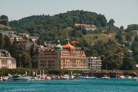A grand building with distinctive architecture featuring an orange roof and the word 'Palace' on it, set against a backdrop of lush green hills. Several boats are docked along a serene body of water in the foreground. The surrounding area includes other buildings, suggesting a picturesque and affluent lakeside town.