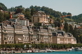 An elegant lakeside town with grand architecture featuring historic hotels and a casino. The buildings are set against a backdrop of lush green hills, and the foreground includes a marina with several yachts docked. The scene is characterized by vibrant greenery and a serene waterfront atmosphere.