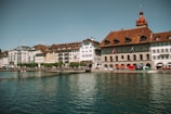 A row of picturesque, multi-story European buildings with varying architectural styles and warm tones lines the edge of a calm body of water. People are strolling along a walkway in front of the buildings, and Swiss flags can be seen hanging from some structures. The sky is clear with a subtle blue hue, suggesting a pleasant day.