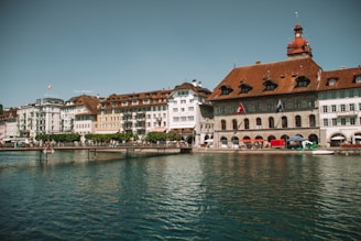 A row of picturesque, multi-story European buildings with varying architectural styles and warm tones lines the edge of a calm body of water. People are strolling along a walkway in front of the buildings, and Swiss flags can be seen hanging from some structures. The sky is clear with a subtle blue hue, suggesting a pleasant day.