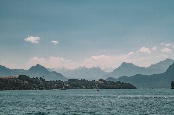 A serene landscape with a large body of water in the foreground, bordered by dense forests and distant mountains under a partly cloudy sky. The scene conveys a sense of tranquility and natural beauty.