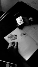 A person is using a smartphone to view an anime character, while sketching the character on a notebook. The scene is captured on a table, with the notebook open displaying partial sketches and the outline of the anime character. The environment appears to be minimally cluttered, emphasizing the artistic focus.