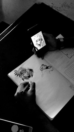 A person is using a smartphone to view an anime character, while sketching the character on a notebook. The scene is captured on a table, with the notebook open displaying partial sketches and the outline of the anime character. The environment appears to be minimally cluttered, emphasizing the artistic focus.