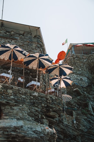 A rustic stone building with several navy and white umbrellas set up on a terrace. Wooden chairs and tables with white tablecloths are arranged under the umbrellas. An Italian flag flutters in the background from the building&rsquo;s upper level. The scene suggests an outdoor seating area or caf&eacute; perched on a cliff or elevated rocky surface.