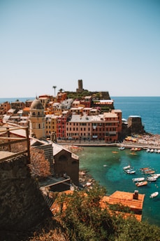 A vibrant coastal village with colorful buildings perched on a cliffside. The clear turquoise sea is filled with small boats, and a crowd of people can be seen enjoying the beach. A historic tower rises prominently above the town, adding to the picturesque scene. The warm sunlight enhances the vivid colors of the buildings and the natural beauty of the landscape.