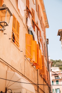 Several vibrant orange shutters are open on the windows of a rustic, light-colored building. Laundry hangs on lines, capturing the essence of a sunlit day. The building appears to be part of a charming street, with a hint of greenery visible in the background.
