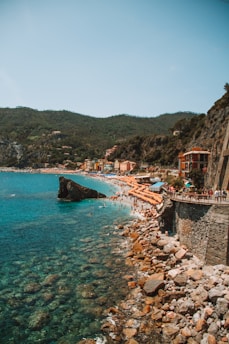 A scenic coastal view featuring a rocky shoreline and a turquoise sea. In the background, colorful buildings are nestled at the base of lush green hills. The beach is populated with numerous orange umbrellas, indicating a bustling seaside destination. People can be seen enjoying the water and sunbathing along the shore.