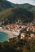 A picturesque coastal town nestled between lush green hills and a serene beachfront. The town features clusters of colorful buildings, primarily with terracotta roofs, lined along the shoreline. The beach is populated with rows of sun loungers and umbrellas, indicating a busy, vibrant tourist spot. The surrounding hills are densely covered with verdant trees and vegetation.