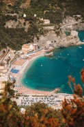 An aerial view of a picturesque coastal area with a long sandy beach lined with rows of orange and blue umbrellas and sun loungers. The beach is adjacent to clear turquoise water, with a few small boats visible in the distance. Surrounding the beach are lush green hills and cliffs, featuring a mix of dense vegetation and some buildings, including a few prominent structures near the shore. The parking area near the beach is filled with numerous cars, suggesting a busy and vibrant atmosphere.