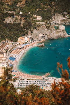 An aerial view of a picturesque coastal area with a long sandy beach lined with rows of orange and blue umbrellas and sun loungers. The beach is adjacent to clear turquoise water, with a few small boats visible in the distance. Surrounding the beach are lush green hills and cliffs, featuring a mix of dense vegetation and some buildings, including a few prominent structures near the shore. The parking area near the beach is filled with numerous cars, suggesting a busy and vibrant atmosphere.