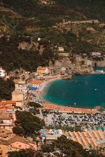 A picturesque coastal town with a sandy beach lined with colorful umbrellas and sunbeds. The clear turquoise water meets the shore, which is crowded with people enjoying the sun. Surrounding the beach are hills covered in lush green vegetation and terraced landscapes. Buildings with bright facades and a parking lot full of cars can be seen in the foreground.