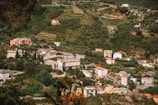 An aerial view of a rural landscape with several buildings of various sizes scattered across a hilly terrain. The area is lush with greenery, including trees and shrubbery, and the buildings are surrounded by gardens and small plots of cultivated land.