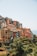 A cluster of colorful Mediterranean-style houses is perched on a hillside, overlooking the sea. The buildings display a variety of vibrant colors like orange, pink, and yellow, and are surrounded by lush greenery. The clear blue sky and distant ocean add to the serene atmosphere.