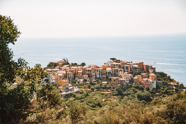 A coastal village with tightly packed, colorful buildings is perched on a cliff overlooking the blue sea. Lush greenery surrounds the village, and the sky is clear with abundant sunlight.