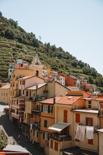 A picturesque hillside village with colorful, stacked buildings featuring red-tiled roofs and vibrant shutters. Lush greenery covers the surrounding hills, indicating a scenic, rural setting. Sunlight casts shadows, enhancing the charm of the architecture.