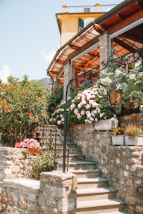 A stone staircase leads up to a house with a rustic design, featuring a wooden pergola and stone pillars. The elegant outdoor area is adorned with lush flowering plants, including pink hydrangeas and various greenery. A vintage lantern hangs from a detailed wrought iron railing, adding to the charm.
