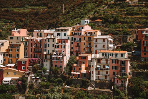 A cluster of vibrant, multicolored buildings stacked closely together on a hillside, surrounded by lush greenery and terraced vineyards. Each building features multiple stories with balconies and shutters, creating a picturesque, quaint atmosphere.