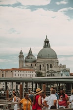 A group of people gathered near a waterfront with a historic architectural structure in the background, featuring large domes and intricate details. The sky is partly cloudy, adding to the grandeur of the scene. The area appears to be a popular tourist spot with a bustling atmosphere.